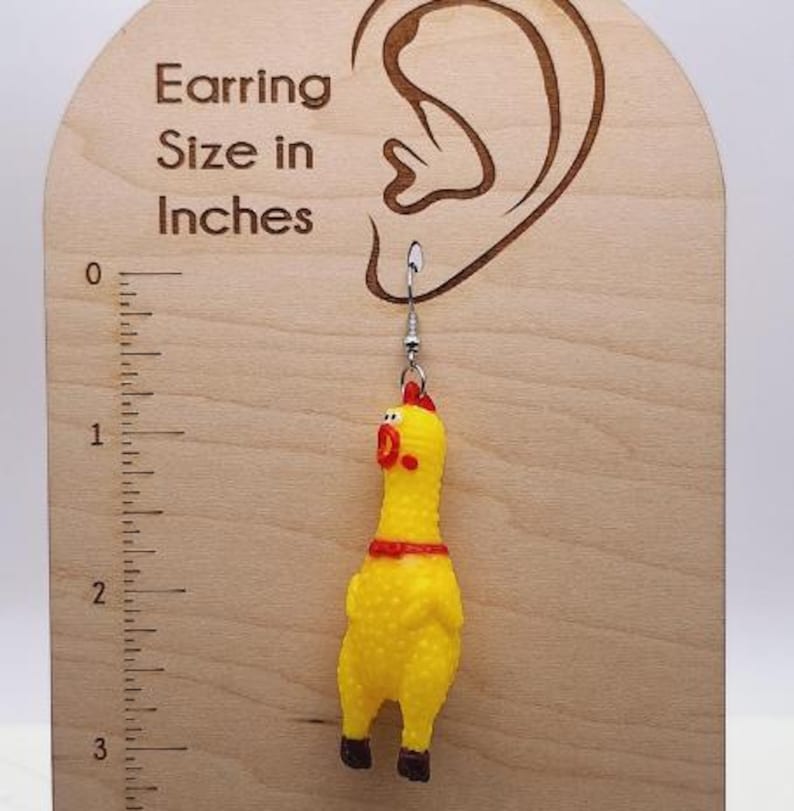 May include: A yellow rubber chicken earring with a red comb and feet, hanging from a silver-toned metal hook. The earring is displayed on a light-colored wooden card featuring an ear outline and a ruler marked in inches, showing the chicken is approximately 2.5 inches long.