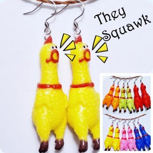 May include: A pair of yellow rubber chicken earrings with red collars and brown feet, featuring silver-toned metal hooks. The text "They Squawk" is printed in black next to yellow graphic sound effect shapes. Additional pairs in various colors are shown.