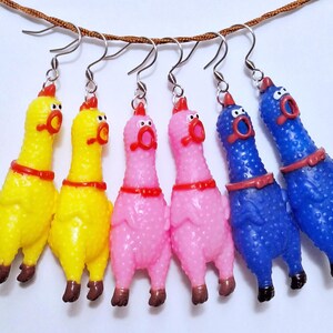 May include: Six dangling earrings shaped like rubber chickens, featuring two yellow, two pink, and two blue figures. Each chicken has a red comb, a wide-open mouth, and a red collar, suspended from silver-toned metal fishhook earring wires.