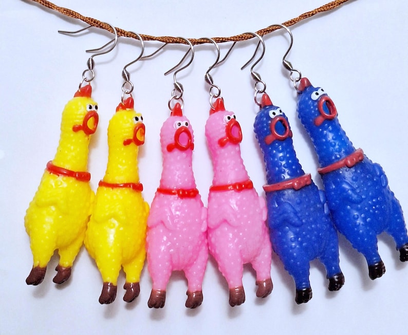 May include: Six dangling earrings shaped like rubber chickens, featuring two yellow, two pink, and two blue figures. Each chicken has a red comb, a wide-open mouth, and a red collar, suspended from silver-toned metal fishhook earring wires.