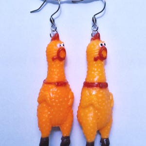 May include: A pair of dangle earrings featuring miniature orange rubber chickens with textured skin, red combs, and dark brown feet. Each chicken has a wide-open mouth and is suspended from a silver-toned metal hook wire for pierced ears.