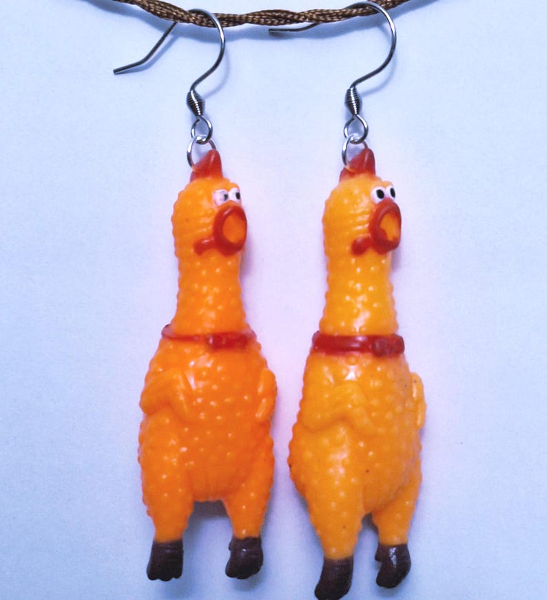 May include: A pair of dangle earrings featuring miniature orange rubber chickens with textured skin, red combs, and dark brown feet. Each chicken has a wide-open mouth and is suspended from a silver-toned metal hook wire for pierced ears.