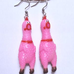 May include: A pair of dangle earrings featuring miniature pink rubber chickens with textured skin, red combs, red beaks, and brown feet. Each chicken has a small red collar and is suspended from a silver-toned metal fishhook earring wire.