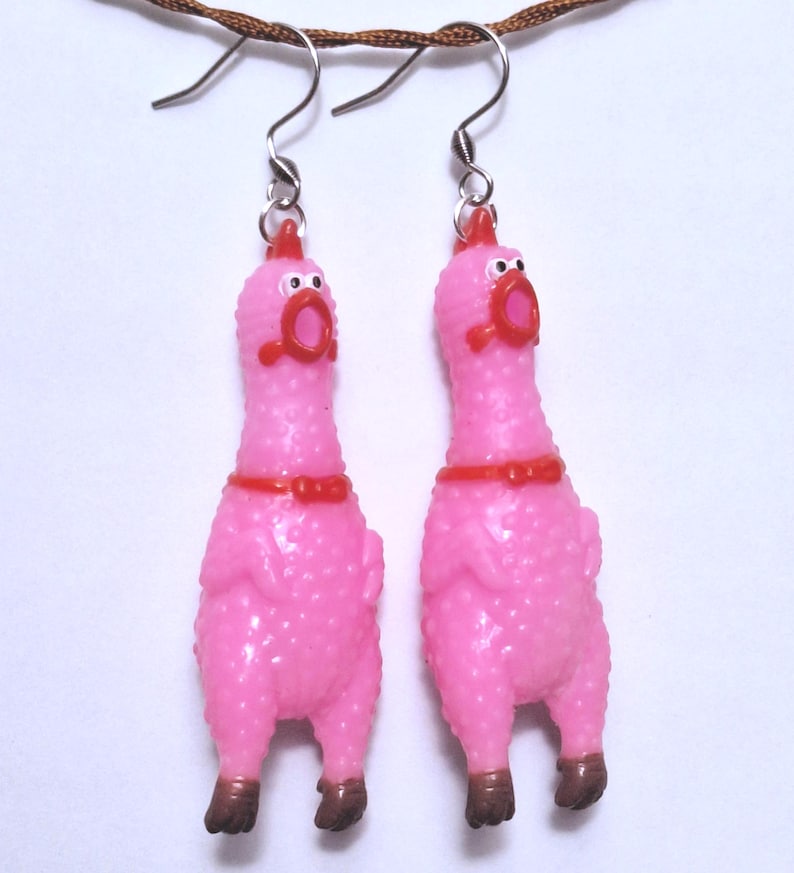 May include: A pair of dangle earrings featuring miniature pink rubber chickens with textured skin, red combs, red beaks, and brown feet. Each chicken has a small red collar and is suspended from a silver-toned metal fishhook earring wire.