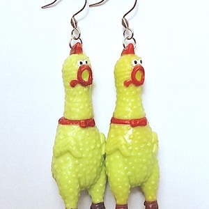 May include: A pair of dangle earrings featuring miniature yellow rubber chickens with textured skin, red combs, red beaks, and brown feet. Each chicken has a wide-open mouth and a small red bow tie around its neck, suspended from silver-toned metal hooks.