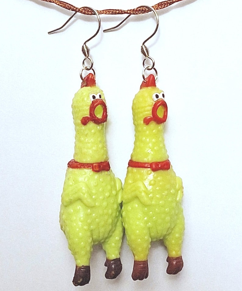 May include: A pair of dangle earrings featuring miniature yellow rubber chickens with textured skin, red combs, red beaks, and brown feet. Each chicken has a wide-open mouth and a small red bow tie around its neck, suspended from silver-toned metal hooks.