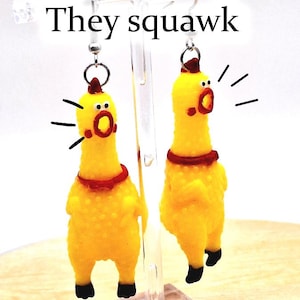 May include: A pair of yellow rubber chicken earrings featuring a textured surface, red neck collars, and black feet. The chickens have open mouths with black lines radiating outward to indicate sound. The text "They squawk" appears above.