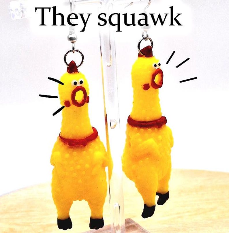 May include: A pair of yellow rubber chicken earrings featuring a textured surface, red neck collars, and black feet. The chickens have open mouths with black lines radiating outward to indicate sound. The text "They squawk" appears above.
