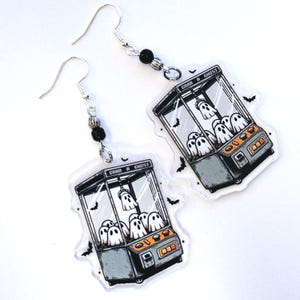 May include: A pair of dangle earrings featuring clear acrylic charms shaped like claw machines filled with white ghosts. The machine displays the text GRAB A GHOST and includes small black bat illustrations. Each earring has a silver hook and black beads.