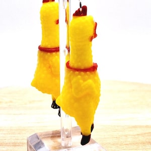 May include: A pair of dangling earrings featuring miniature yellow rubber chickens with textured skin, red neck details, and dark brown tail feathers. The chickens are suspended from a clear acrylic display stand, showcasing their bright yellow color.