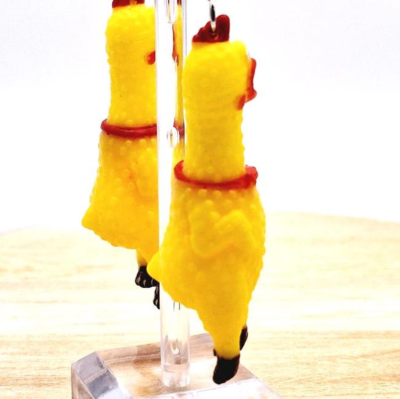 May include: A pair of dangling earrings featuring miniature yellow rubber chickens with textured skin, red neck details, and dark brown tail feathers. The chickens are suspended from a clear acrylic display stand, showcasing their bright yellow color.