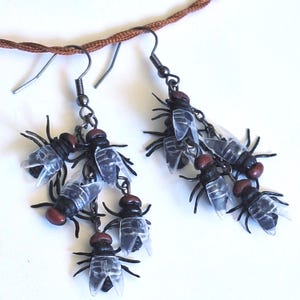 May include: A pair of dangle earrings featuring multiple miniature black fly charms with reddish-brown heads and translucent plastic wings. The flies are attached to dark metal chains suspended from standard metal earring hooks.