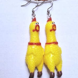 May include: A pair of dangling earrings featuring miniature yellow rubber chickens with red combs, red beaks, and brown feet. The chickens have wide, surprised expressions with white eyes and black pupils, suspended from silver-toned metal earring hooks.
