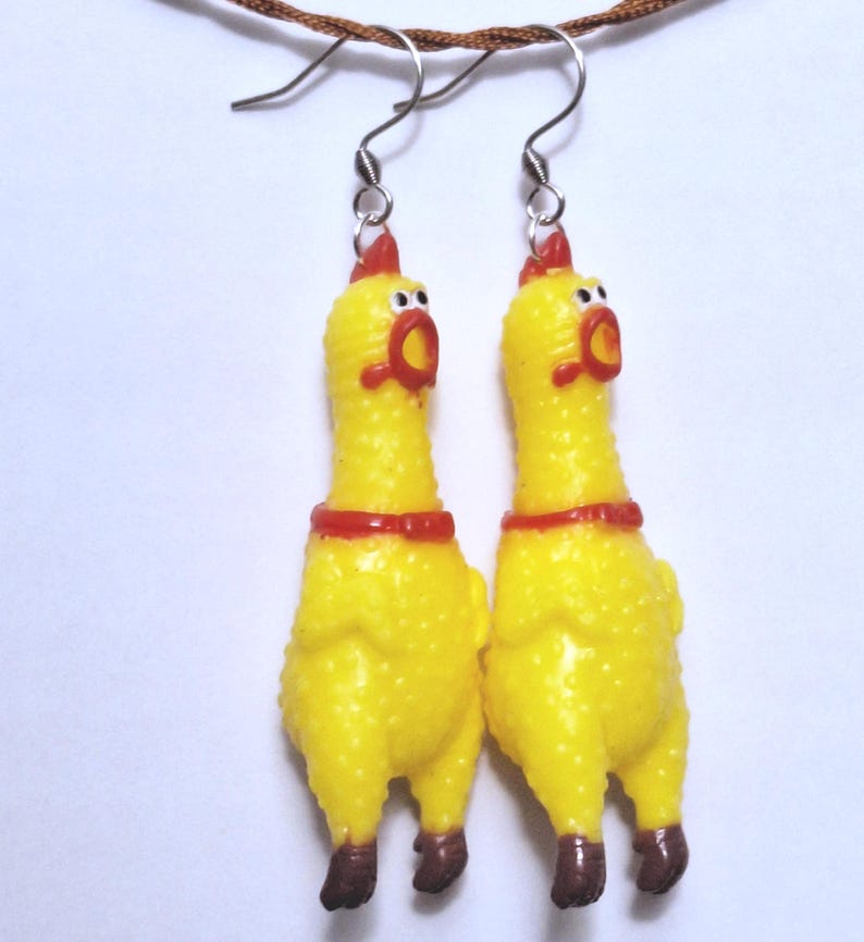 May include: A pair of dangling earrings featuring miniature yellow rubber chickens with red combs, red beaks, and brown feet. The chickens have wide, surprised expressions with white eyes and black pupils, suspended from silver-toned metal earring hooks.