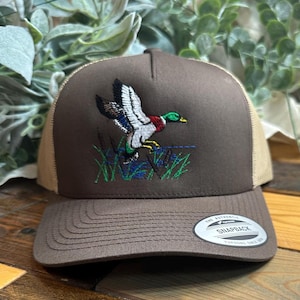 May include: A brown and tan trucker hat with an embroidered image of a mallard duck in flight. The hat has a mesh back and a snap closure. The text "The Authentic Snapback" is printed on a silver sticker on the back of the hat.