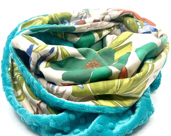 Pode incluir: Um cachecol infinito turquesa com um padrão floral. O cachecol apresenta uma camada externa turquesa macia e texturizada e uma camada interna vibrante com desenhos florais verdes, brancos e laranja. Projetado para ser usado ao redor do pescoço.