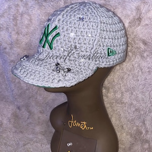 May include: A gray crocheted baseball cap with a green New York Yankees logo and silver embellishments. The cap is on a brown mannequin head.