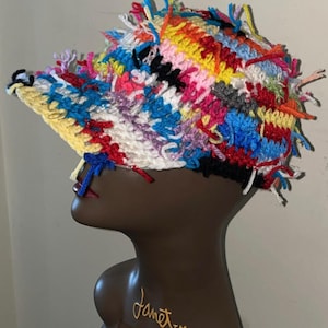 May include: A colorful crocheted hat with a long brim and a messy, frayed texture. The hat is made with a variety of colors, including red, blue, yellow, green, and pink.