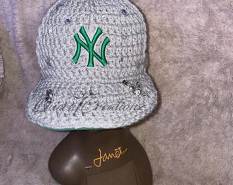 May include: Gray crocheted baseball cap with a green embroidered New York Yankees logo. The cap has a wide brim and is decorated with silver embellishments.