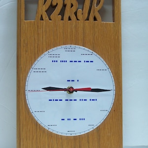 May include: A wooden clock with a white face and black hands. The clock face has morse code for the letters K2RJk.