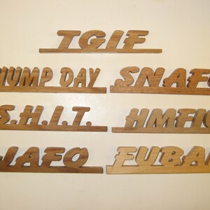 Puede incluir: Un conjunto de ocho letreros de madera con varios acrónimos y frases, incluyendo "TGIF", "Hump Day", "SNAFU", "S.H.I.T.", "HMFFIC", "JAFO" y "FUBAR".