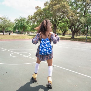 May include: A person wearing a blue floral dress and white knee-high socks is roller skating on a basketball court. They are wearing a blue drawstring backpack with a roller skate pattern.