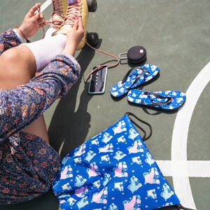 May include: A blue drawstring bag with a roller skate pattern. The bag is lying on a green surface with a white line. There are also a pair of flip flops, a phone, and a speaker on the surface.