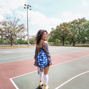May include: A woman wearing a floral dress and white socks is roller skating on a basketball court. She is carrying a blue drawstring bag with a roller skate pattern.