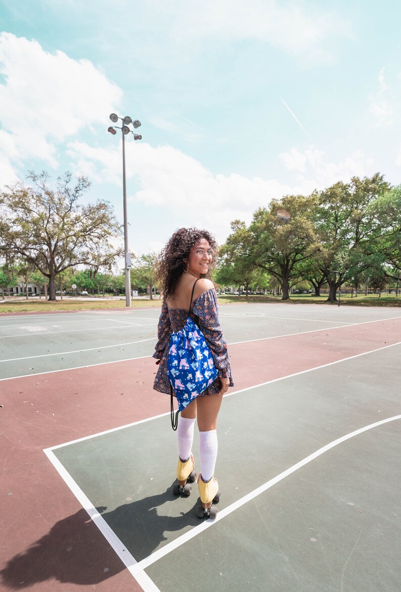 May include: A woman wearing a floral dress and white socks is roller skating on a basketball court. She is carrying a blue drawstring bag with a roller skate pattern.