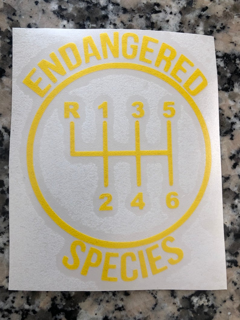 May include: A yellow and white sticker with the text "Endangered Species" in a circle around a gear shift pattern. The gear shift pattern has the numbers 1, 2, 3, 4, 5, and 6, and the letter "R" for reverse.