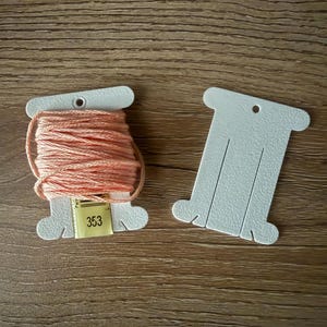 May include: Two white cardboard bobbins for embroidery thread. One bobbin is wrapped with peach-coloured thread and features a small yellow label with the number 353 printed in black. The other bobbin is empty and plain white with a hole at the top.