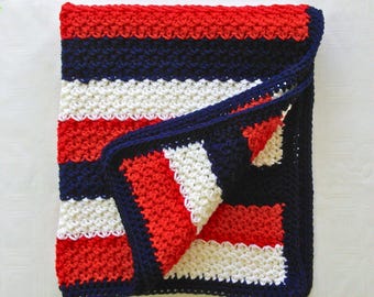 May include: A folded, handmade crochet blanket featuring alternating stripes of red, navy blue, and white. The blanket has a navy blue border and is made with a textured stitch. The blanket is folded over, revealing the interior pattern.