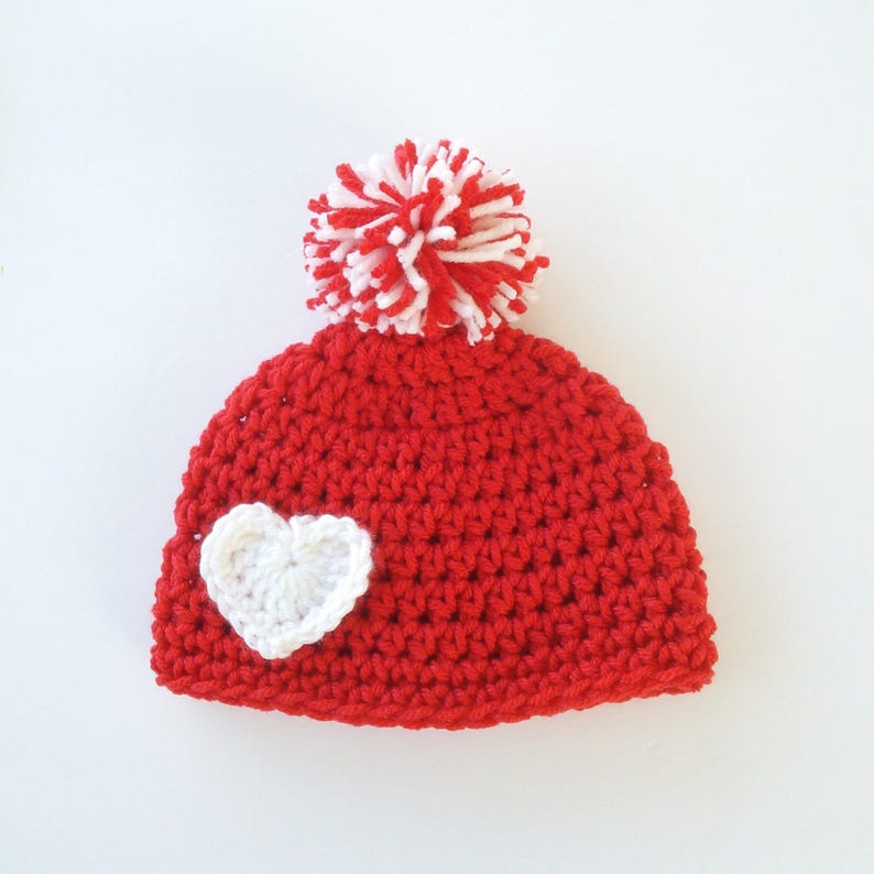 May include: Red crocheted beanie with a white heart and a white and red pom pom on top.