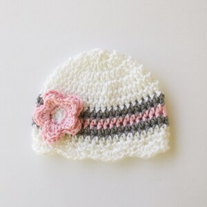 May include: A white crocheted baby hat with a pink flower and a striped band of gray and pink. The hat is handmade and has a scalloped edge. The flower is centered on the hat.