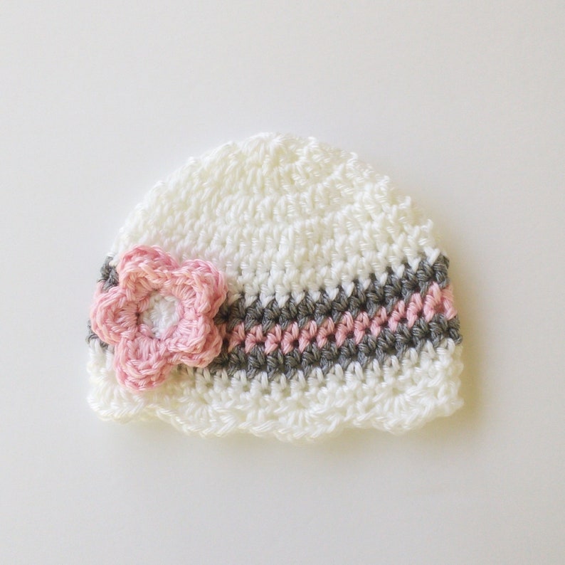 May include: A white crocheted baby hat with a pink flower and a striped band of gray and pink. The hat is handmade and has a scalloped edge. The flower is centered on the hat.