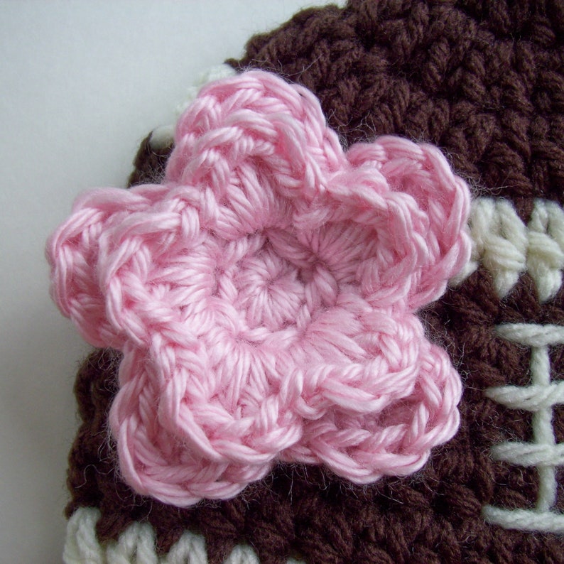 Peut inclure: Gros plan sur un article crochet&eacute; &agrave; la main avec une grande fleur rose. La fleur est centr&eacute;e sur un fond crochet&eacute; marron et blanc. L'article semble &ecirc;tre un chapeau ou un bonnet.