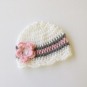 May include: A white crocheted baby hat with a pink flower and a striped design in gray and pink. The hat has a scalloped edge and is handmade. This baby hat is a perfect accessory for newborns.