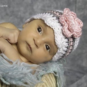 May include: A newborn baby wearing a handmade crochet hat. The hat is white, gray, and pink, with a large pink flower on the side. The baby is lying on a soft, blue blanket.