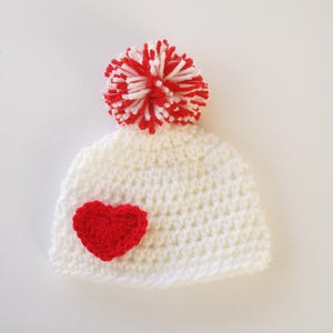 May include: White crocheted beanie with a red heart applique and a red and white pom pom on top.