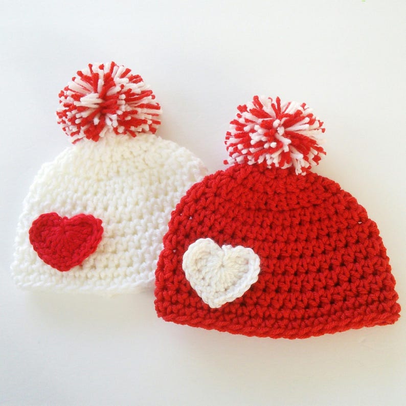 May include: Two crocheted hats with pom-poms. One hat is white with a red heart and the other is red with a white heart.
