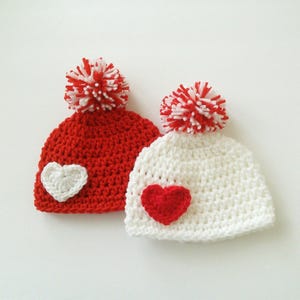 May include: Two crocheted hats with pom-poms. One hat is red with a white heart and the other is white with a red heart.
