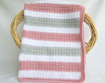 Puede incluir: Una manta de bebé a rayas hecha a mano en una cesta. La manta presenta rayas horizontales alternas en blanco, rosa empolvado y verde claro. Hecha de un material suave y texturizado.
