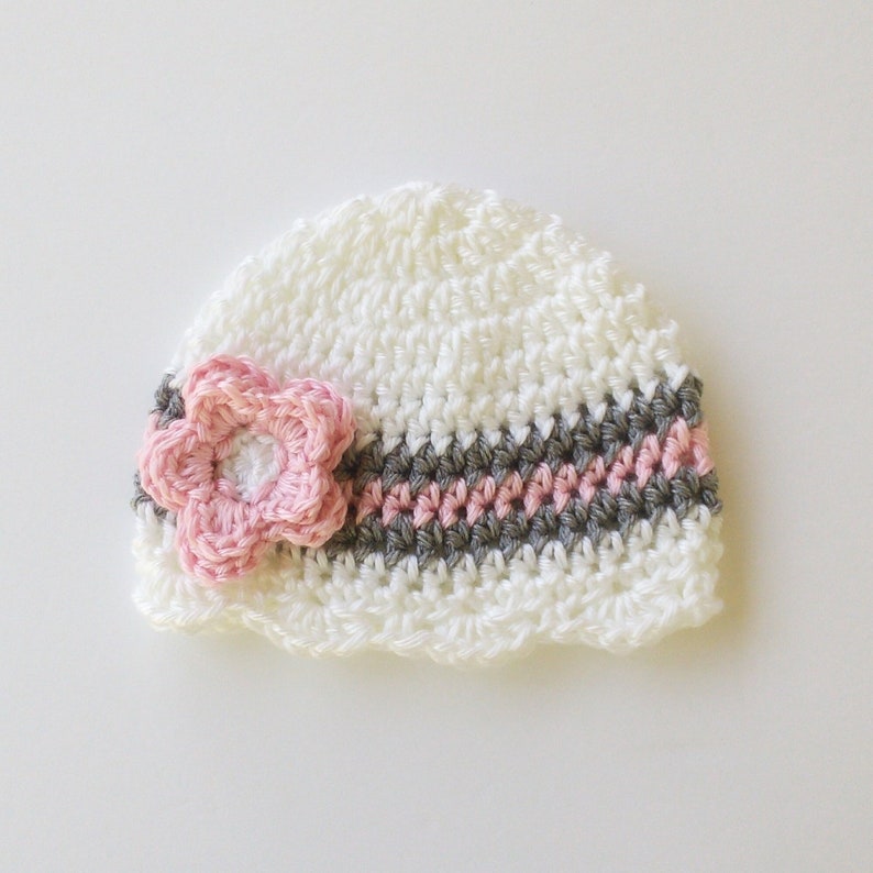 May include: A white crocheted baby hat with a pink flower and a striped band of gray and pink. The hat is handmade and has a scalloped edge. The flower is a 3D design.