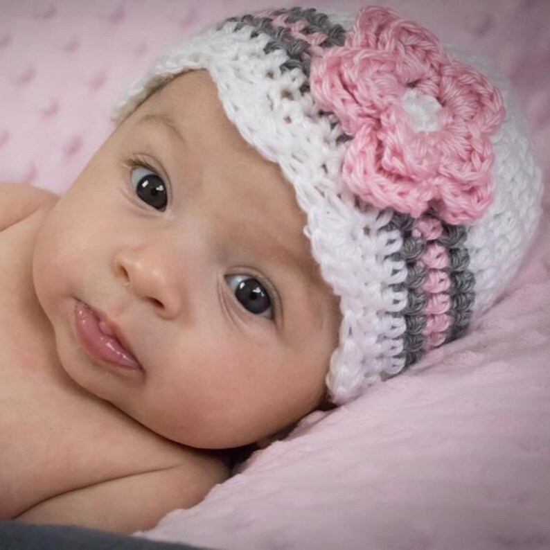 May include: A baby wearing a white, gray, and pink crocheted hat with a large pink flower on the side. The baby is lying on a pink blanket.