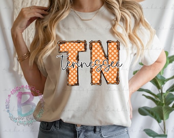 May include: Cream-colored t-shirt featuring the word "Tennessee" in a bold, orange polka-dot design with black outlines. The letters "TN" are prominent, with the state name written below in a cursive font. The shirt has short sleeves.