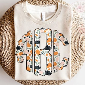 May include: A cream-colored short-sleeve t-shirt featuring a large, stylized monogram of the letters ABC. The letters are filled with a repeating pattern of orange, black, and light pink pumpkins, green leaves, and small foliage on a white background.