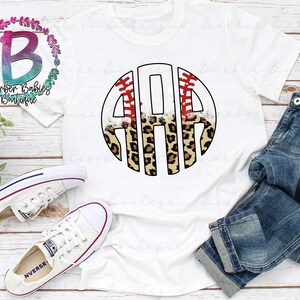 May include: White t-shirt featuring a baseball-themed monogram design with the letters "ARA" in a circular frame. The design incorporates baseball stitching, leopard print, and the text "Barber Babies Boutique". The shirt is paired with blue jeans and white sneakers.