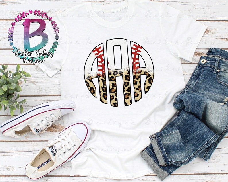 May include: White t-shirt featuring a baseball-themed monogram design with the letters "ARA" in a circular frame. The design incorporates baseball stitching, leopard print, and the text "Barber Babies Boutique". The shirt is paired with blue jeans and white sneakers.
