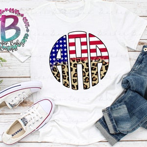 May include: White t-shirt featuring a circular design with the letters "AMA" in red and white stripes, blue stars, and leopard print. The shirt is paired with blue jeans and white sneakers. The text "Barber Babies Boutique" is also visible.