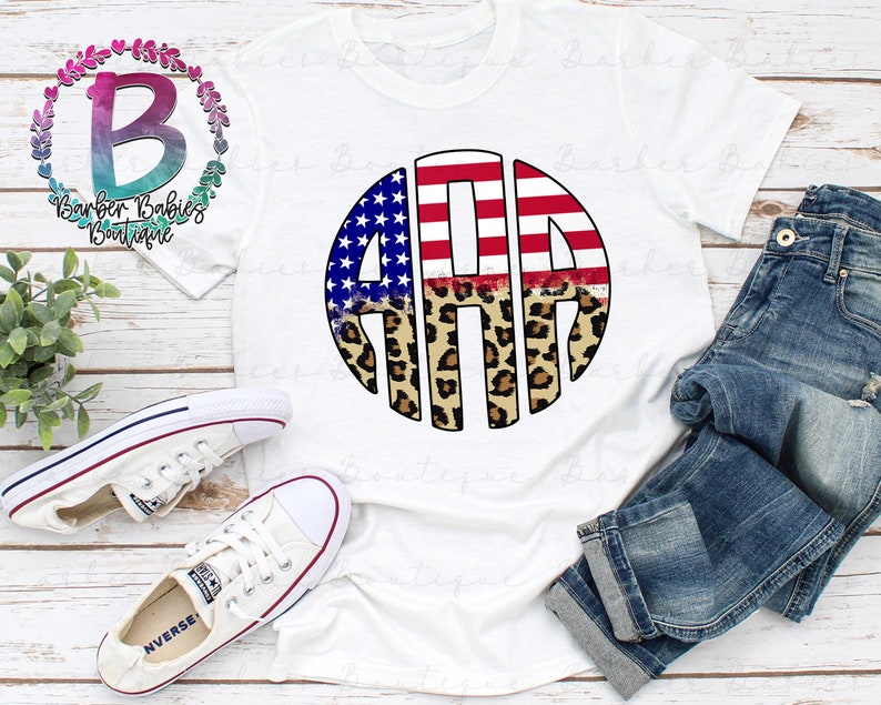 May include: White t-shirt featuring a circular design with the letters "AMA" in red and white stripes, blue stars, and leopard print. The shirt is paired with blue jeans and white sneakers. The text "Barber Babies Boutique" is also visible.